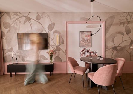 Stylowa część jadalna apartamentu z różowymi aksamitnymi krzesłami, okrągłym ciemnym stołem, roślinną tapetą, telewizorem, lampą i rozmazanym gościem.