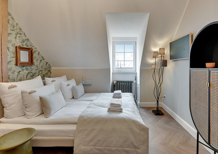 Przytulny pokój na poddaszu ze skośnym sufitem, wzorzystą ścianą, białą pościelą, złożonymi ręcznikami i szafą z rattanu