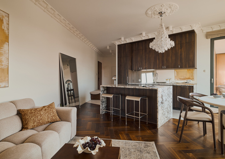 Przestronny apartament z wygodną sofą, marmurową wyspą kuchenną, ciemnymi drewnianymi szafkami, kryształowym żyrandolem i stołem przy balkonie