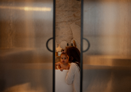 Ciepła łazienka hotelowa z marmurem, mama kąpie dziecko w wolnostojącej wannie widocznej przez mleczne drzwi.