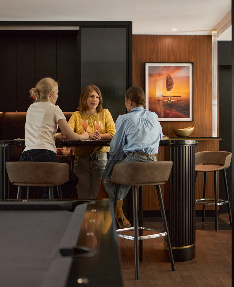 Trzy kobiety rozmawiają przy barowym stoliku, popijając koktajle w eleganckim salonie z bilardem