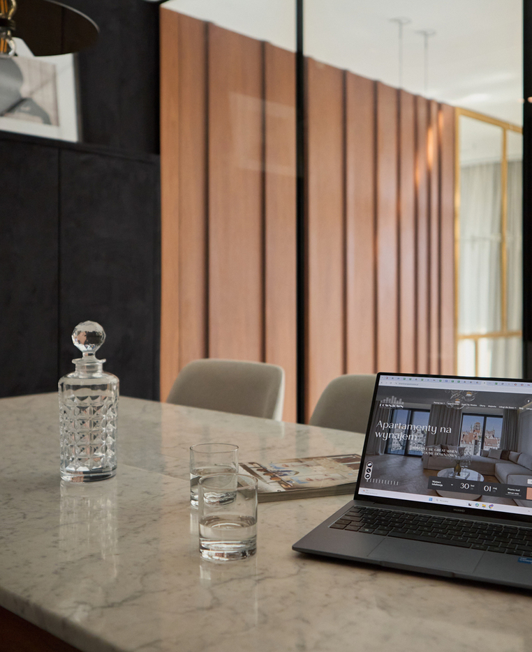 Stół z marmurowym blatem jako kącik do pracy z laptopem pokazującym apartamenty na wynajem, kryształowy dekanter, szklanki i ciepłe drewniane panele.