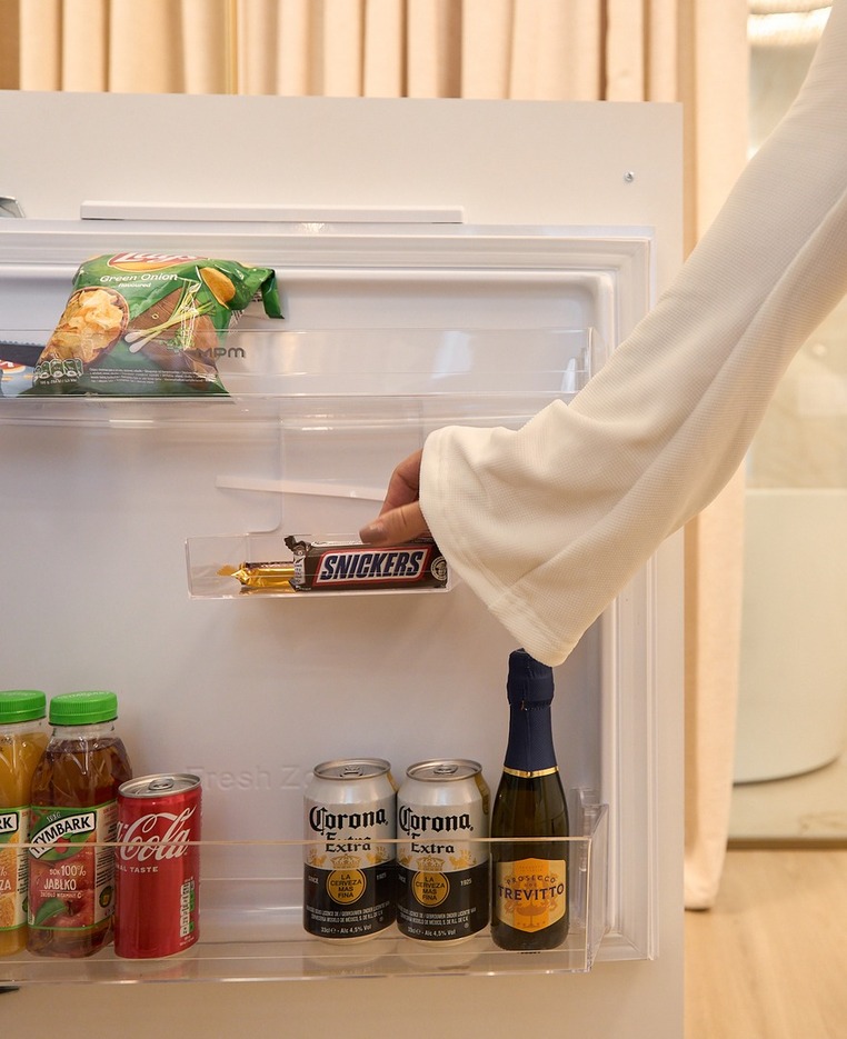 Kompaktowa minibarowa lodówka w pokoju z przekąskami, sokami, Coca-Colą, piwem Corona i prosecco; ręka sięga po Snickersa.