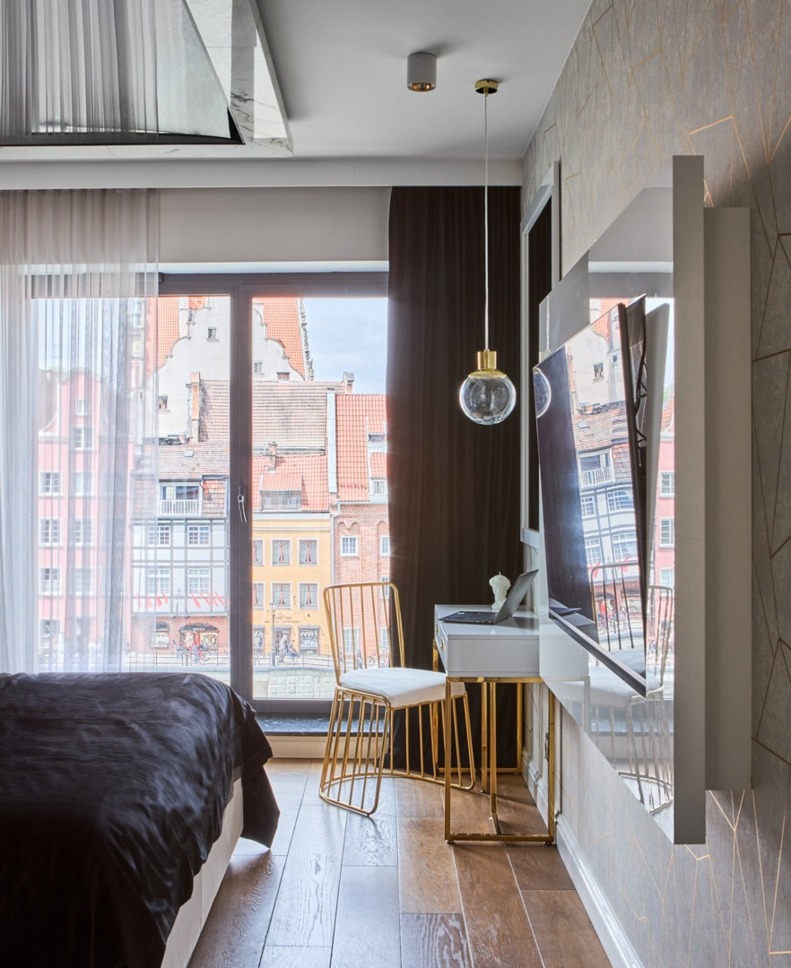 Nowoczesny pokój hotelowy z podwójnym łóżkiem w czarnej pościeli, wiszącymi lampami, biurkiem ze złotym krzesłem i widokiem na kolorowe kamienice