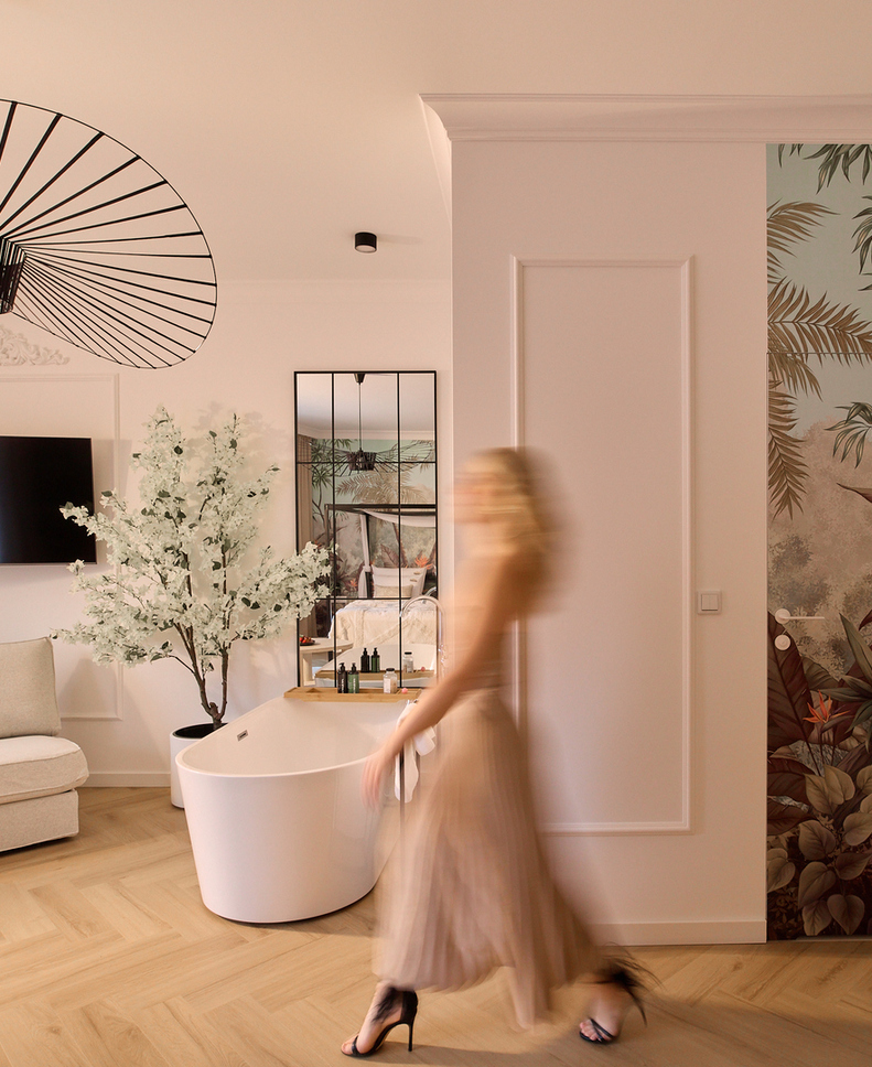 Przestronny nowoczesny apartament z wolnostojącą wanną, fotelem, telewizorem, tropikalnym muralem i roślinami; gość w ruchu.