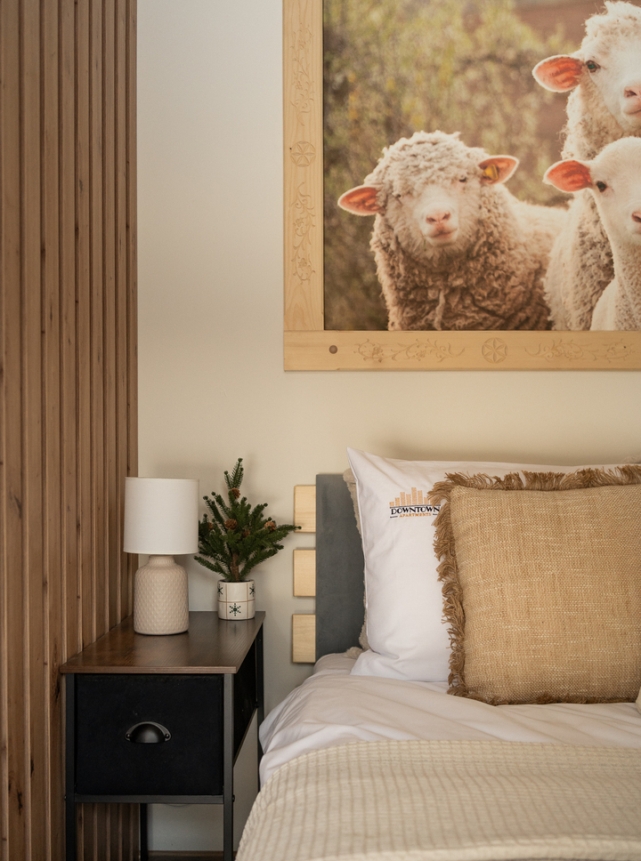 Przytulny pokój z fakturowaną pościelą i poduszką z frędzlami, lampką nocną, małą choinką na stoliku, obrazem owiec i drewnianą lamperią.