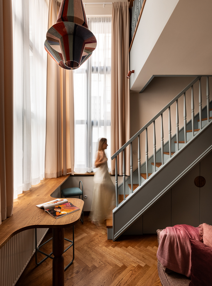 Dwupoziomowy apartament z antresolą, wysokimi oknami, drewnianym biurkiem, jodełkową podłogą, kolorową lampą i gościem wchodzącym po schodach.