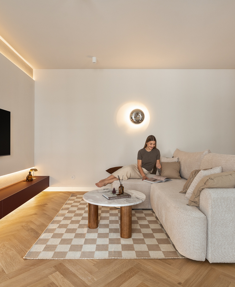 Nowoczesny salon apartamentu z miękką narożną sofą, marmurowym stolikiem, telewizorem i ciepłym oświetleniem; gość czyta.