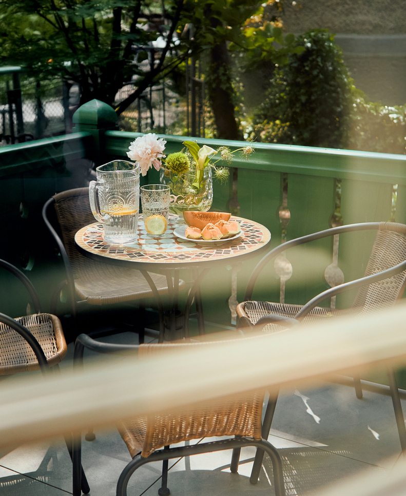 Przytulny balkon hotelowy z mozaikowym stolikiem, wiklinowymi fotelami, dzbankiem wody z cytryną, kawałkami melona i kwiatami.