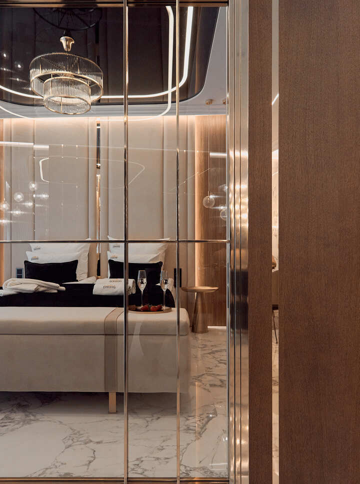 Luksusowy apartament przez przeszkloną przegrodę — łóżko king-size z czarnymi poduszkami, ławeczka z szampanem i truskawkami, żyrandol, marmurowa podłoga, ciepłe oświetlenie