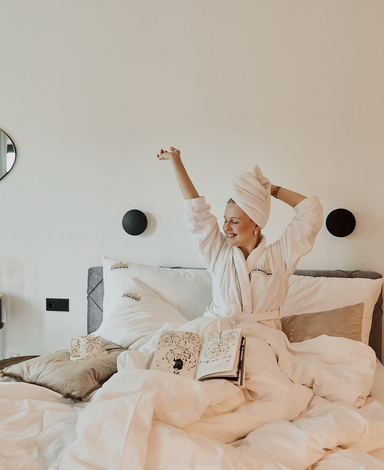 Gość w szlafroku z turbanem na głowie rozciąga się na łóżku z białą, puszystą pościelą, otwartą książką, lampkami nocnymi i biurkiem