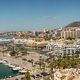 Marina con yacht, passeggiata fiancheggiata da palme, hotel e appartamenti sul lungomare con costa e montagne sullo sfondo