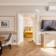 Suite luminosa con zona giorno, tavolo da pranzo, sedie in velluto, TV a parete e parquet; porte aperte rivelano letto dorato e lampadario.