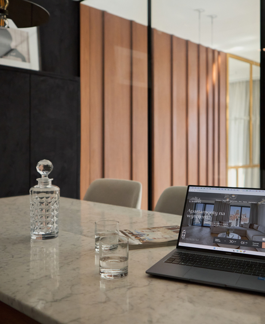 Marmurowy stół w nowoczesnym apartamencie, kryształowa karafka, szklanki i laptop z ofertą wynajmu.