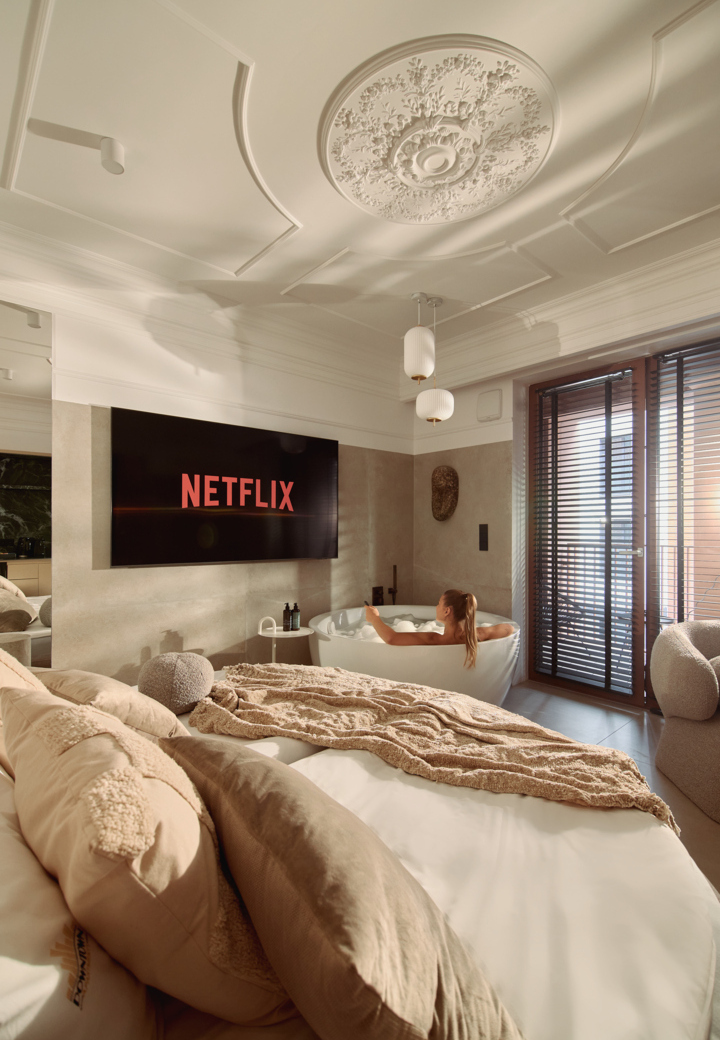 Apartament z łóżkiem i wolnostojącą wanną, gdzie gość ogląda Netflix na telewizorze; ozdobna rozetka sufitowa, lampy i balkon.