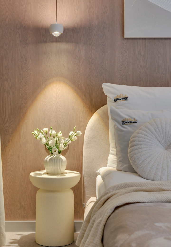 Przytulne nocne miejsce w hotelu: lampa wisząca, biały wazon z kwiatami na stoliku, miękkie poduszki i pled na tapicerowanym łóżku.