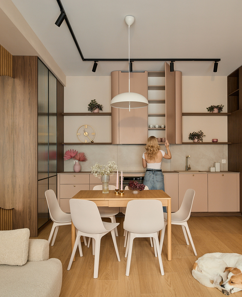 Kuchnia w aneksie z pastelowymi szafkami, drewnianymi akcentami, stołem na 6 osób i lampą wiszącą; gość sięga do szafki, pies śpi.