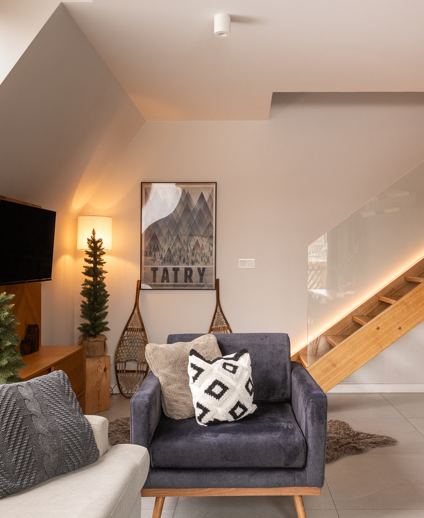 Przytulny salon na poddaszu z welurowym fotelem, kanapą, drewnianymi schodami z LED i oknem z widokiem gór