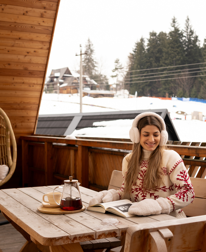 Przytulny balkon w chacie z drewnianym stołem i huśtawką koszową; gość w nausznikach czyta, herbata i widok na zaśnieżone stoki.