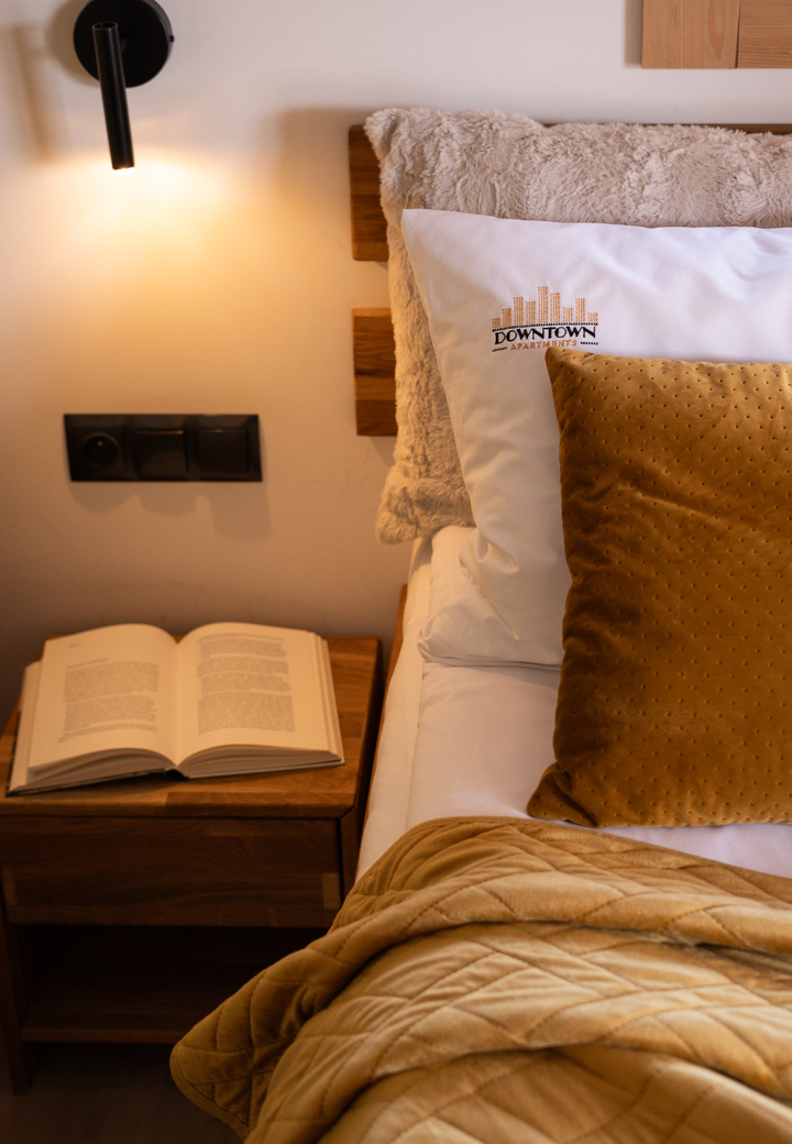 Przytulne miejsce przy łóżku z miękkim zagłówkiem, poduszką z haftem 'Downtown', złotym poduszkiem i kocem, lampką i otwartą książką