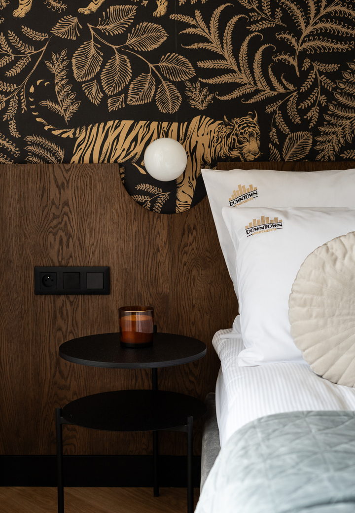 Przyłóżkowy kącik w stylu butikowym: złota tapeta z tygrysem, drewniany zagłówek, kulista lampa, okrągły stolik z bursztynowym kieliszkiem i poduszki z logo.