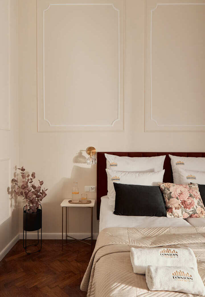 Eleganckie łóżko dwuosobowe z puszystą pościelą, haftowanymi poduszkami i ręcznikami, kwiatowy jasiek, karafka i kinkiet