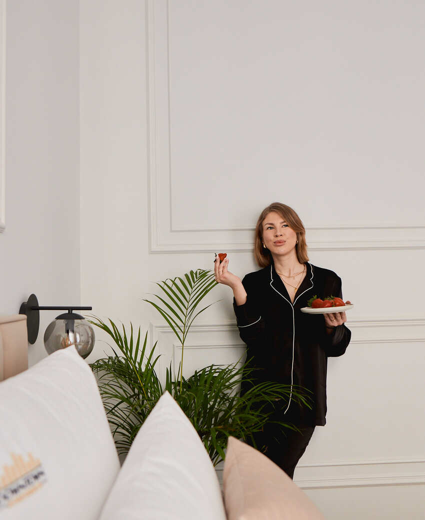 Gość w czarnych piżamach je truskawki w jasnym pokoju hotelowym z białymi ozdobnymi panelami, palmą w doniczce i miękkimi poduszkami.