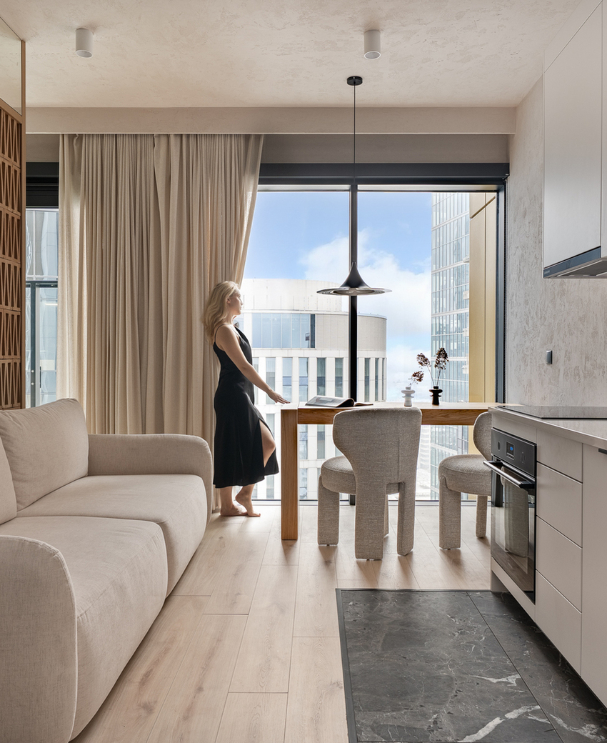 Nowoczesny apartament typu studio z aneksem, beżową sofą, stołem przy dużym oknie i osobą w czarnej sukience patrzącą na miasto.