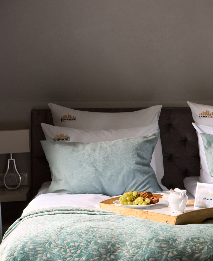 Przytulne łóżko dwuosobowe z turkusowymi poduszkami i wzorzystą narzutą; taca ze śniadaniem, winogronami i rogalikami przy oknie.