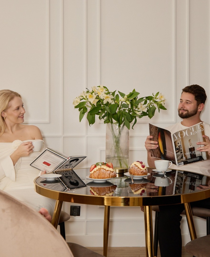 Para przy śniadaniu w eleganckiej sali hotelowej z kawą, croissantami z owocami, magazynami i bukietem lilii.