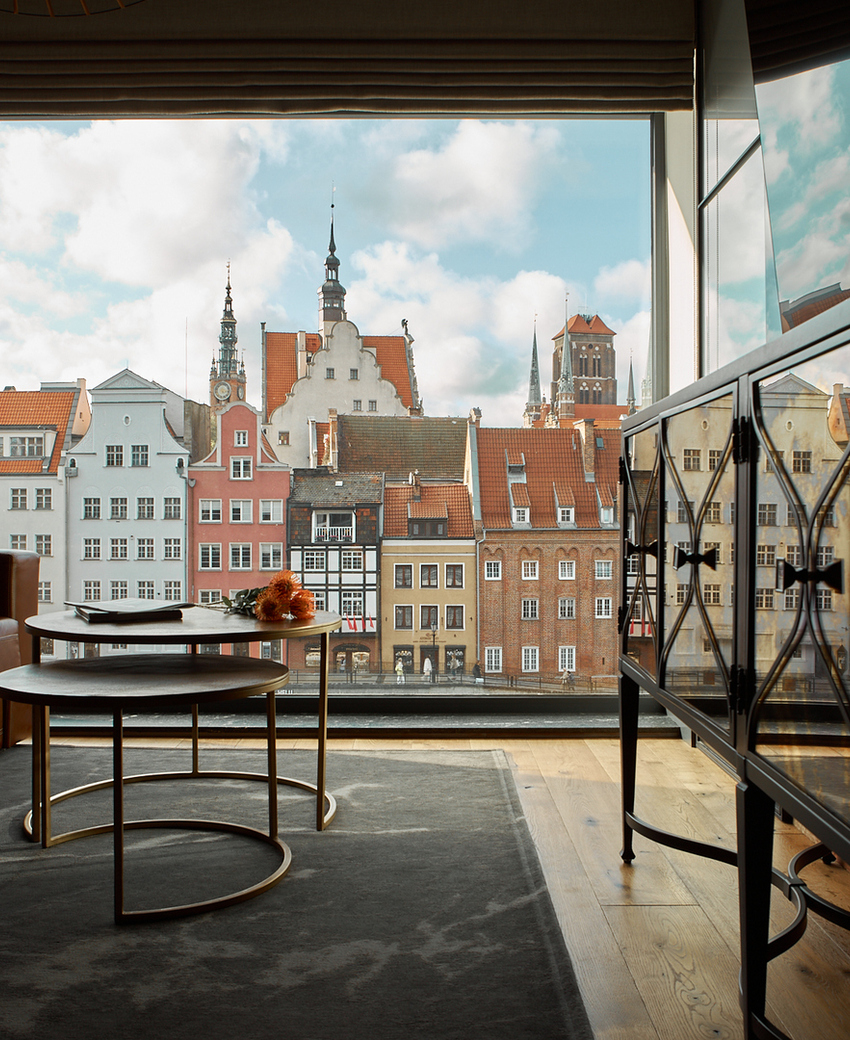 Salon w apartamencie z skórzaną sofą, stolikami i dużym oknem z widokiem na kolorowe kamienice i wieże kościołów.