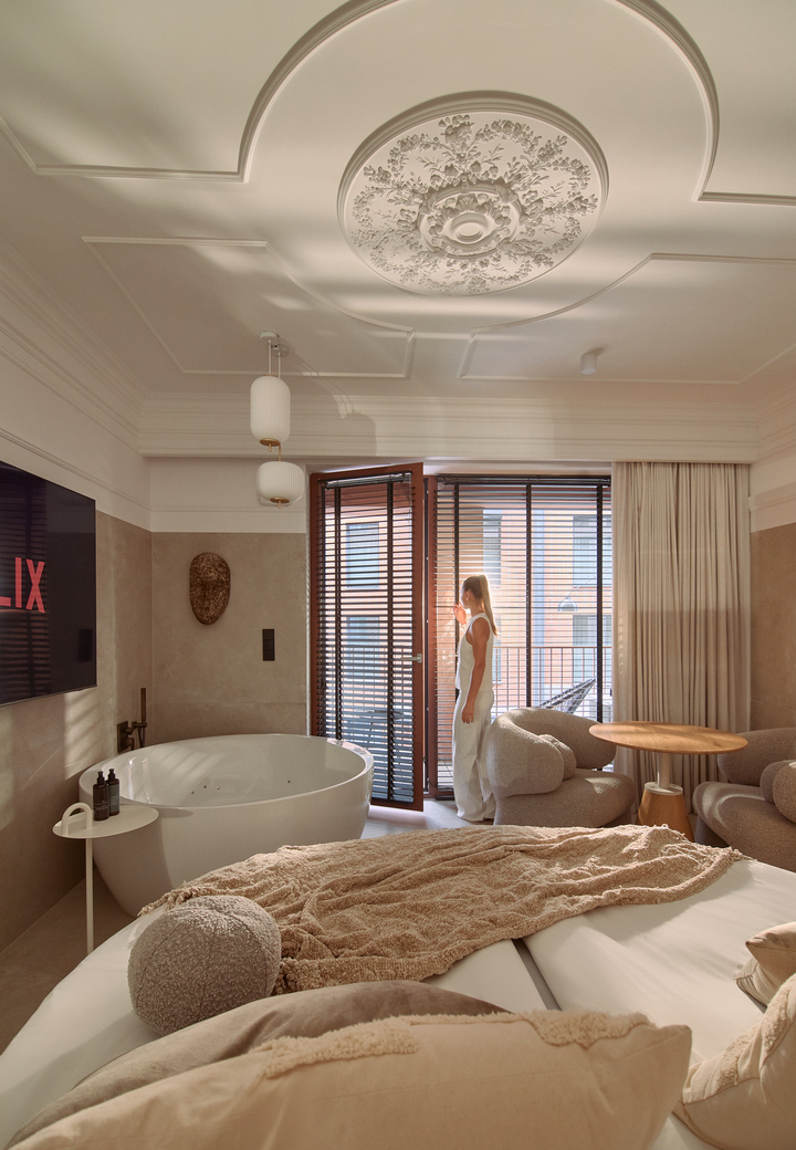 Butikowy apartament z ozdobnym medalionem sufitowym, wolnostojącą wanną, przytulnym łóżkiem i kącikiem wypoczynkowym, kobieta przy drzwiach balkonu