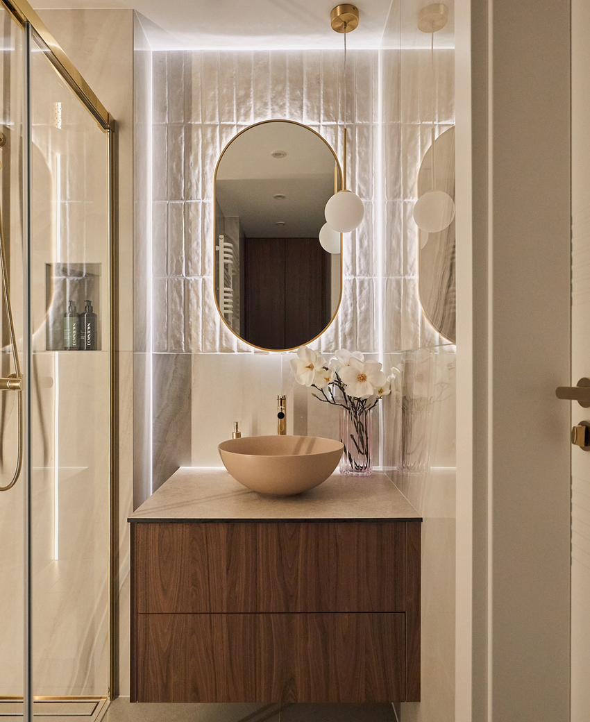 Nowoczesna łazienka z przeszklonym prysznicem i złotymi armaturami, umywalką nablatową na drewnianej szafce, podświetlonym owalnym lustrem i orchideą