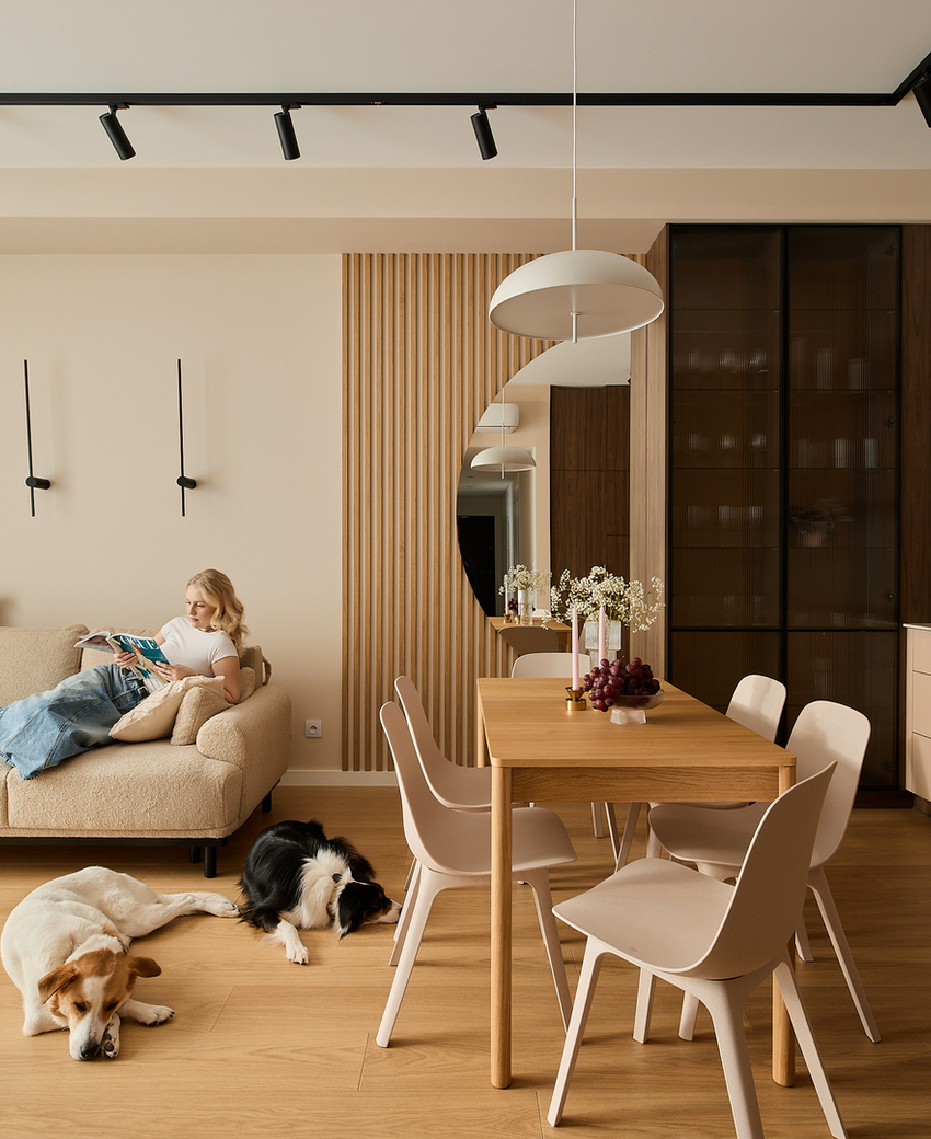 Przestronny salon z beżową sofą, gość czyta, dwa psy odpoczywają na drewnianej podłodze, stół jadalniany i nowoczesny aneks kuchenny