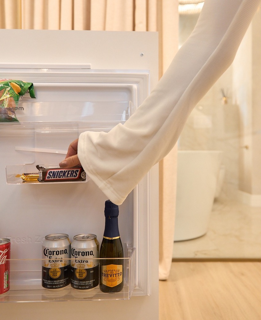 Kompaktowa minibarowa lodówka w pokoju z przekąskami, sokami, Coca-Colą, piwem Corona i prosecco; ręka sięga po Snickersa.