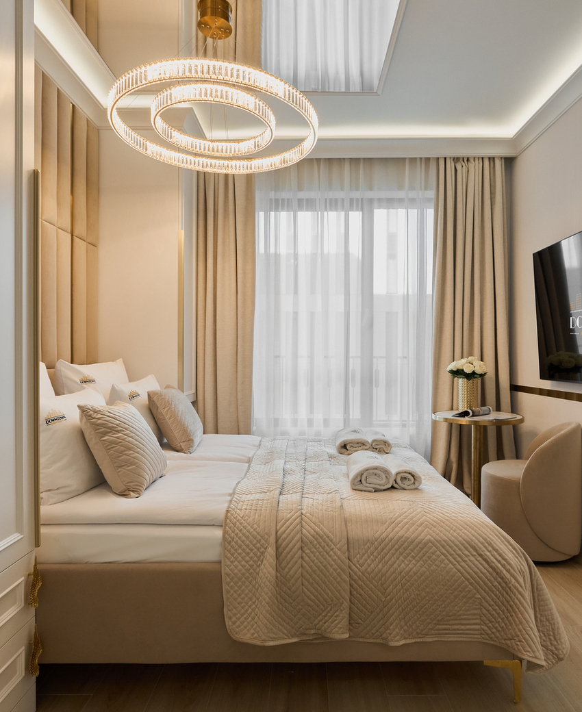 Elegancki pokój hotelowy w stonowanych tonach z dużym łóżkiem, pikowaną pościelą, okrągłym żyrandolem, TV i zwiniętymi ręcznikami.