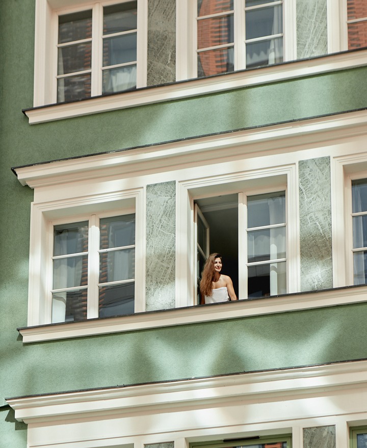 Gość w otwartym oknie butikowego hotelu nad parterową kawiarnią, zielona elewacja, poranne słońce i relaksująca atmosfera