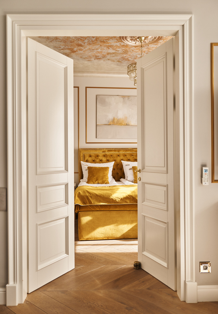 Podwójne drzwi do słonecznej, eleganckiej sypialni z pikowanym złotym łóżkiem, żyrandolem, ozdobnym sufitem i parkietem w jodełkę