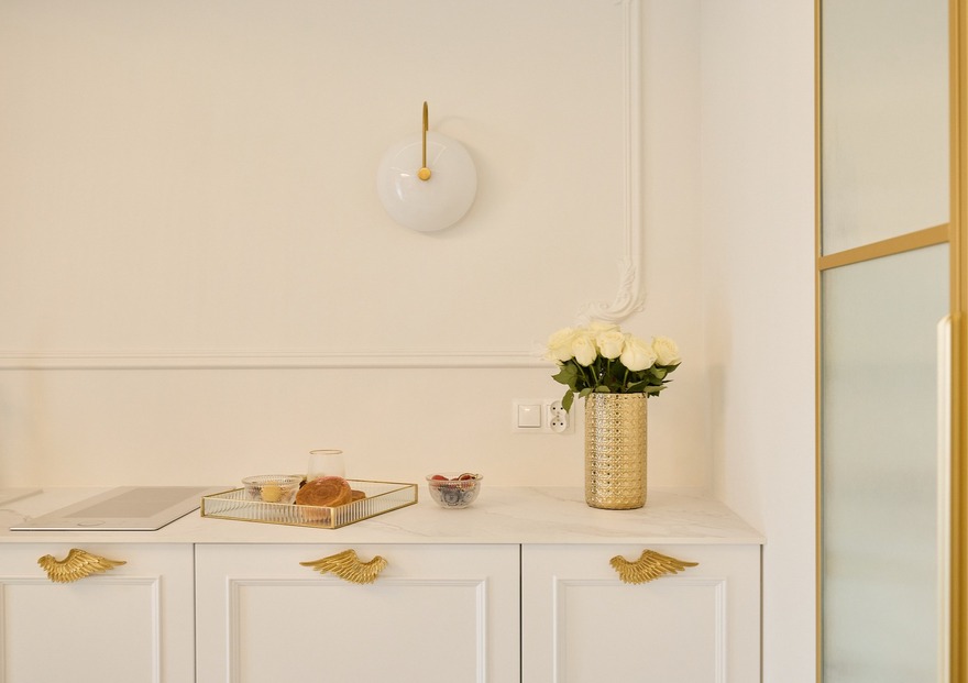 Butikowy aneks kuchenny z marmurowym blatem, złotymi uchwytami-skrzydłami, złotym wazonem z białymi różami i tacą z wypiekami