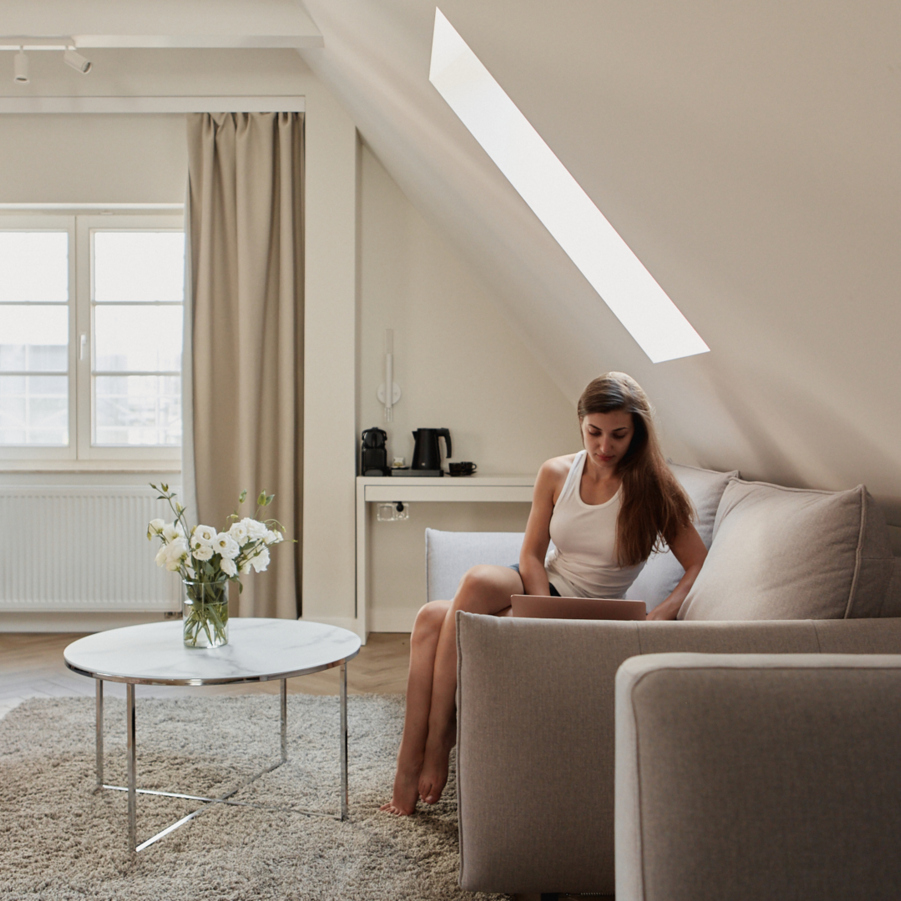 Poddaszowa suite ze skośnym oknem dachowym, stonowane wnętrze, sofa i łóżko, marmurowy stolik z kwiatami, ekspres i gość przy laptopie.