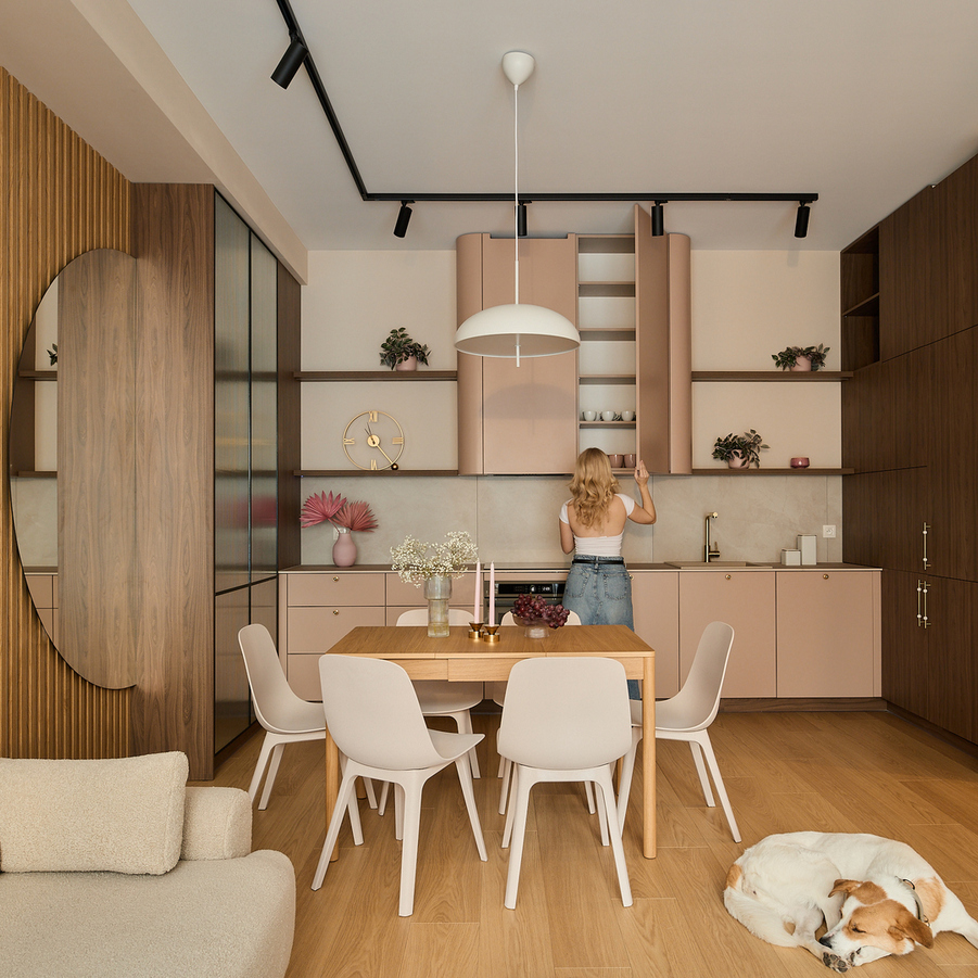 Kuchnia w aneksie z pastelowymi szafkami, drewnianymi akcentami, stołem na 6 osób i lampą wiszącą; gość sięga do szafki, pies śpi.