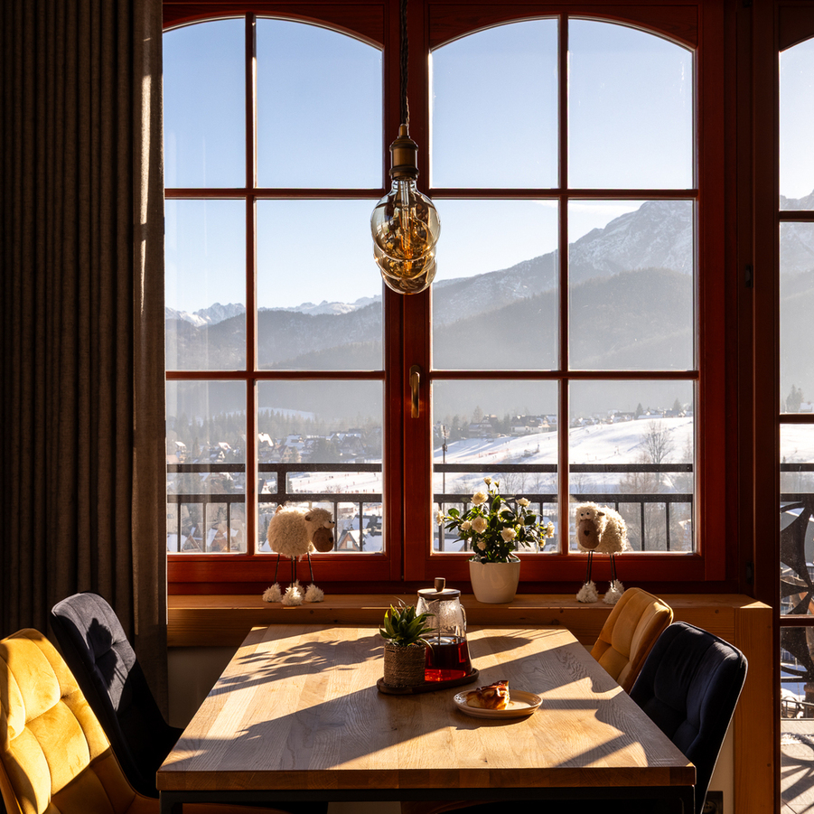 Przytulny stół przy łukowych oknach z widokiem na zaśnieżone góry, drewniany blat, wisząca lampa, kwiaty i herbata.