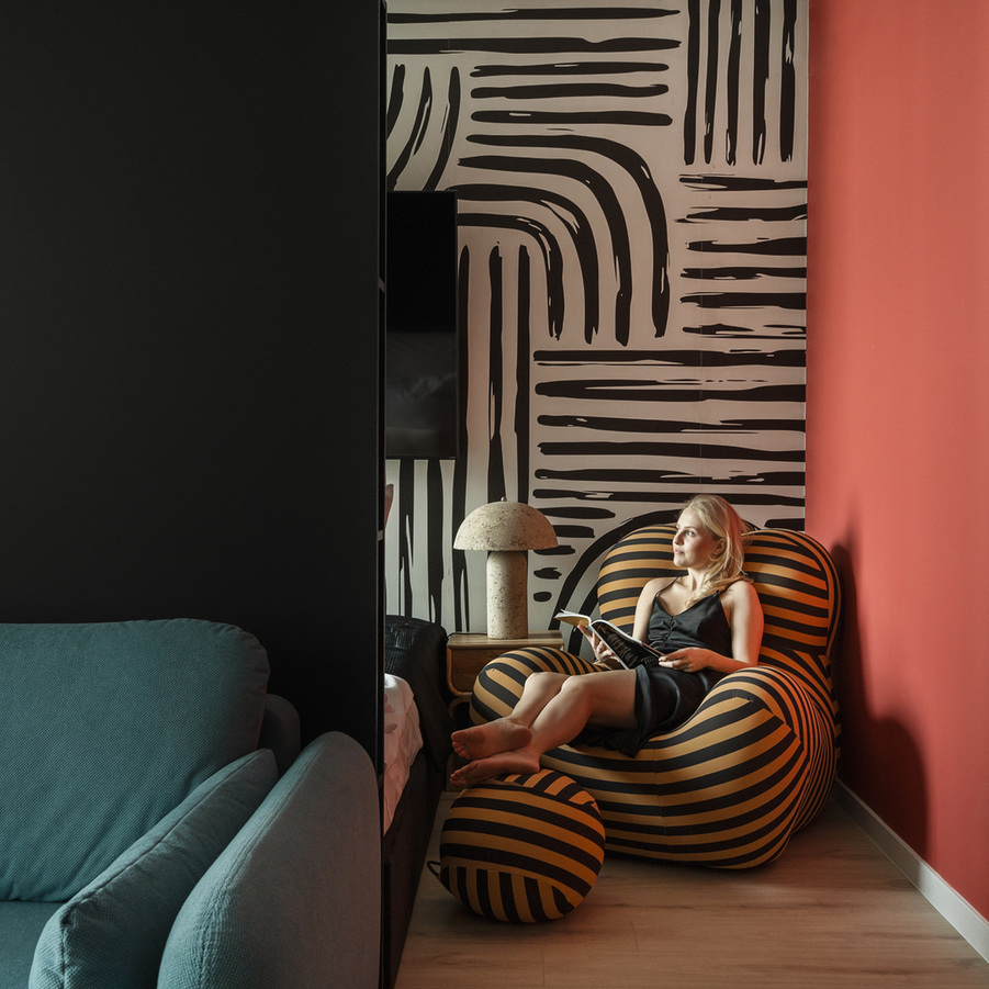 Nowoczesny hotelowy kącik do czytania z pasiastym fotelem i podnóżkiem, graficzną tapetą, lampą w kształcie grzyba i gościem czytającym.