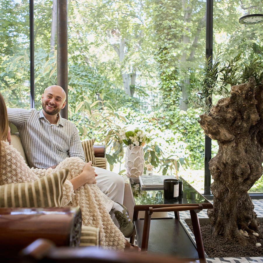 Para relaksuje w hotelowym lounge ze szklanymi ścianami i widokiem na ogród, przytulna pasowana sofa, kawa na stoliku i ozdobny pień.