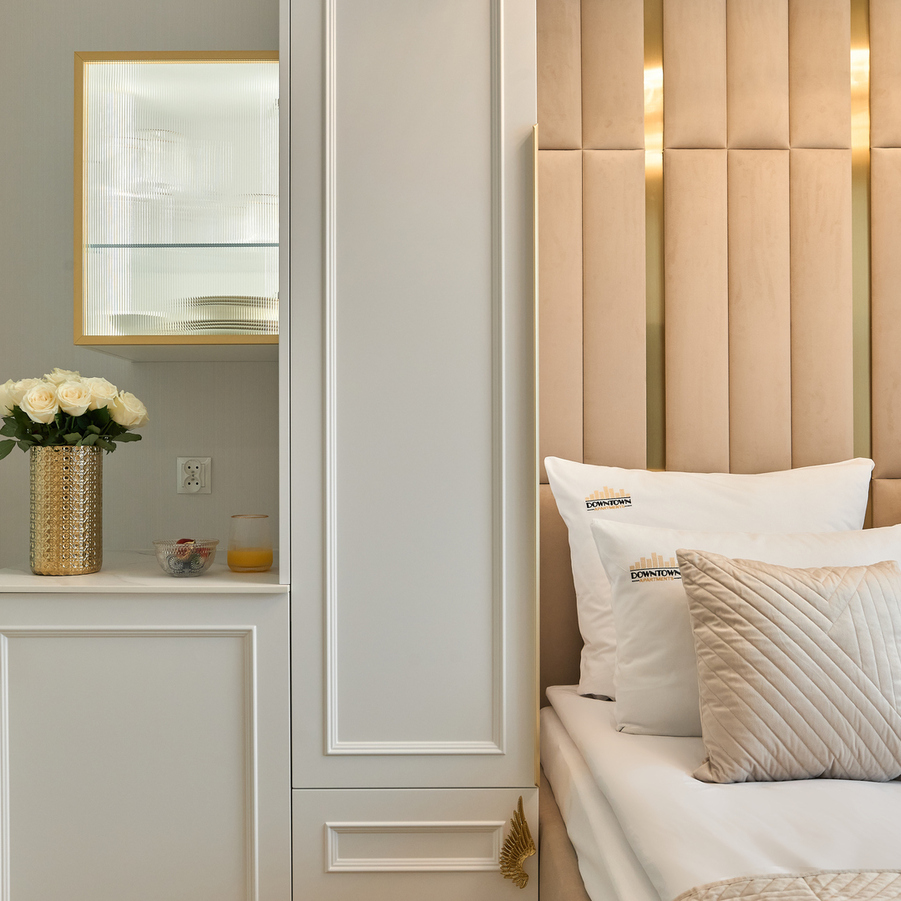 Przytulne łóżka z wysokim pikowanym beżowym zagłówkiem, białe poduszki z logo, ozdobna pikowana poduszka, złote akcenty i wazon z białymi różami