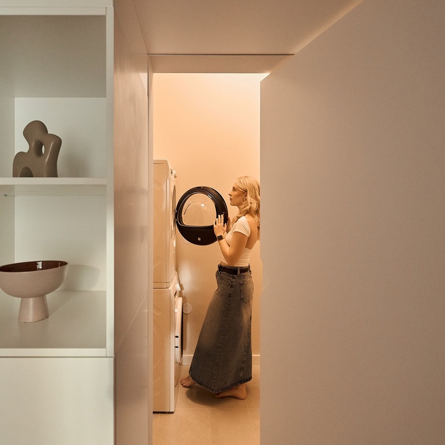 Gość wkłada pranie do pionowej pralko-suszarki w ciepłej, minimalistycznej pralni hotelowej z dekoracyjną półką.