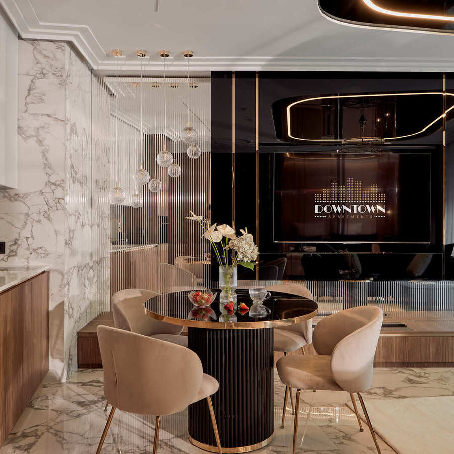 Jadalnia w luksusowym apartamencie z marmurową podłogą, drewnianym aneksem, okrągłym czarnym stołem ze złotą krawędzią i beżowymi fotelami.