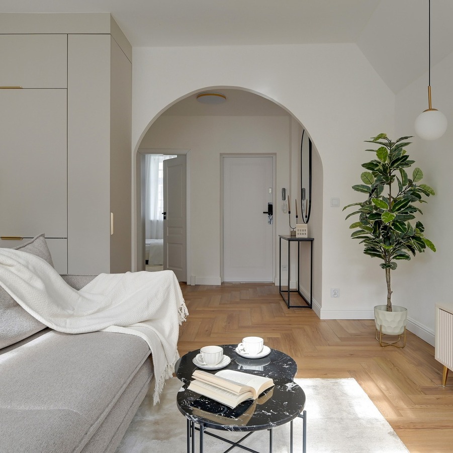 Jasny, nowoczesny salon w apartamencie z łukowym przejściem, zabudowaną kuchnią, szarą sofą i stolikiem z filiżankami.