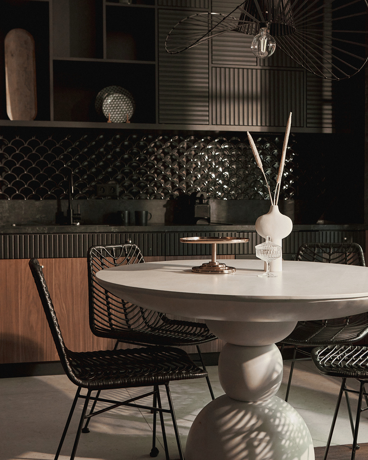 Nowoczesny kącik jadalny z okrągłym stołem na cokole, plecionymi czarnymi krzesłami, ciemnym fakturowanym panelem i rzeźbiarską lampą