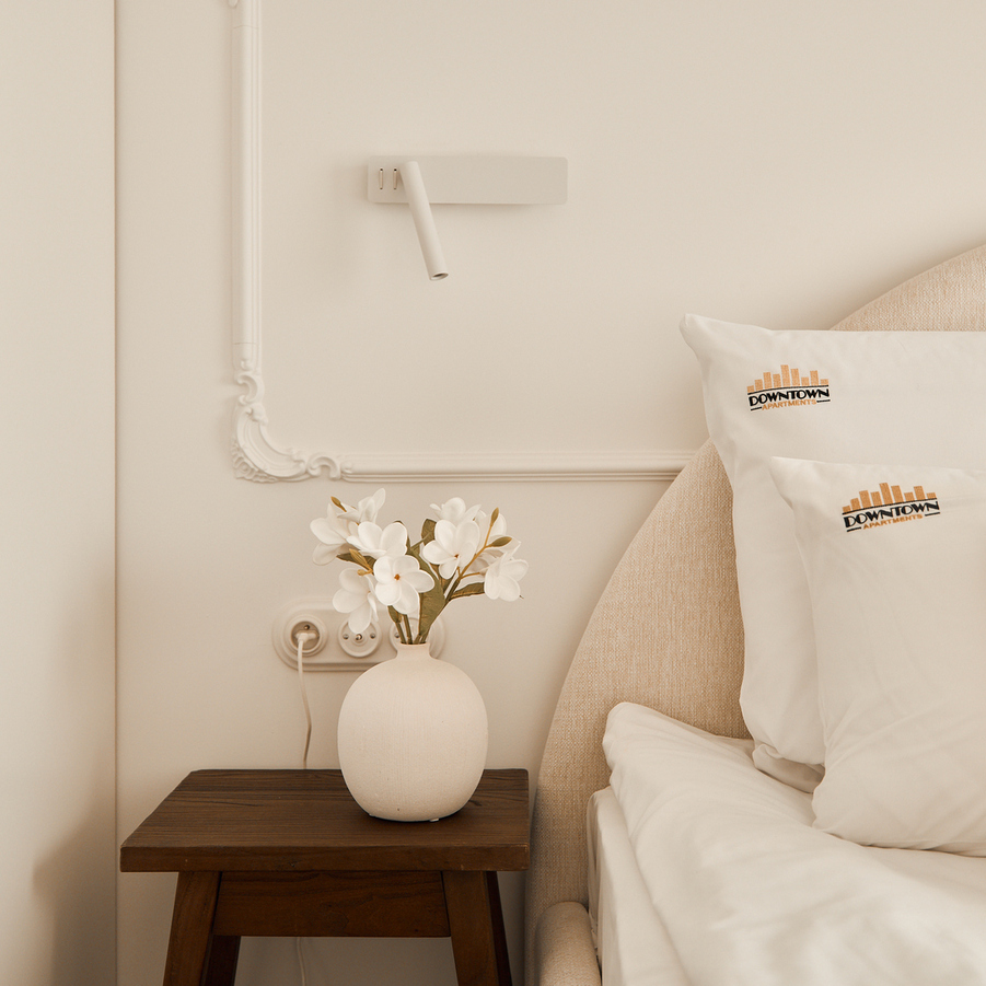 Przytulny kąt pokoju z tapicerowanym zagłówkiem, białą pościelą, poduszkami z logo, stolikiem nocnym z wazonem i lampką do czytania